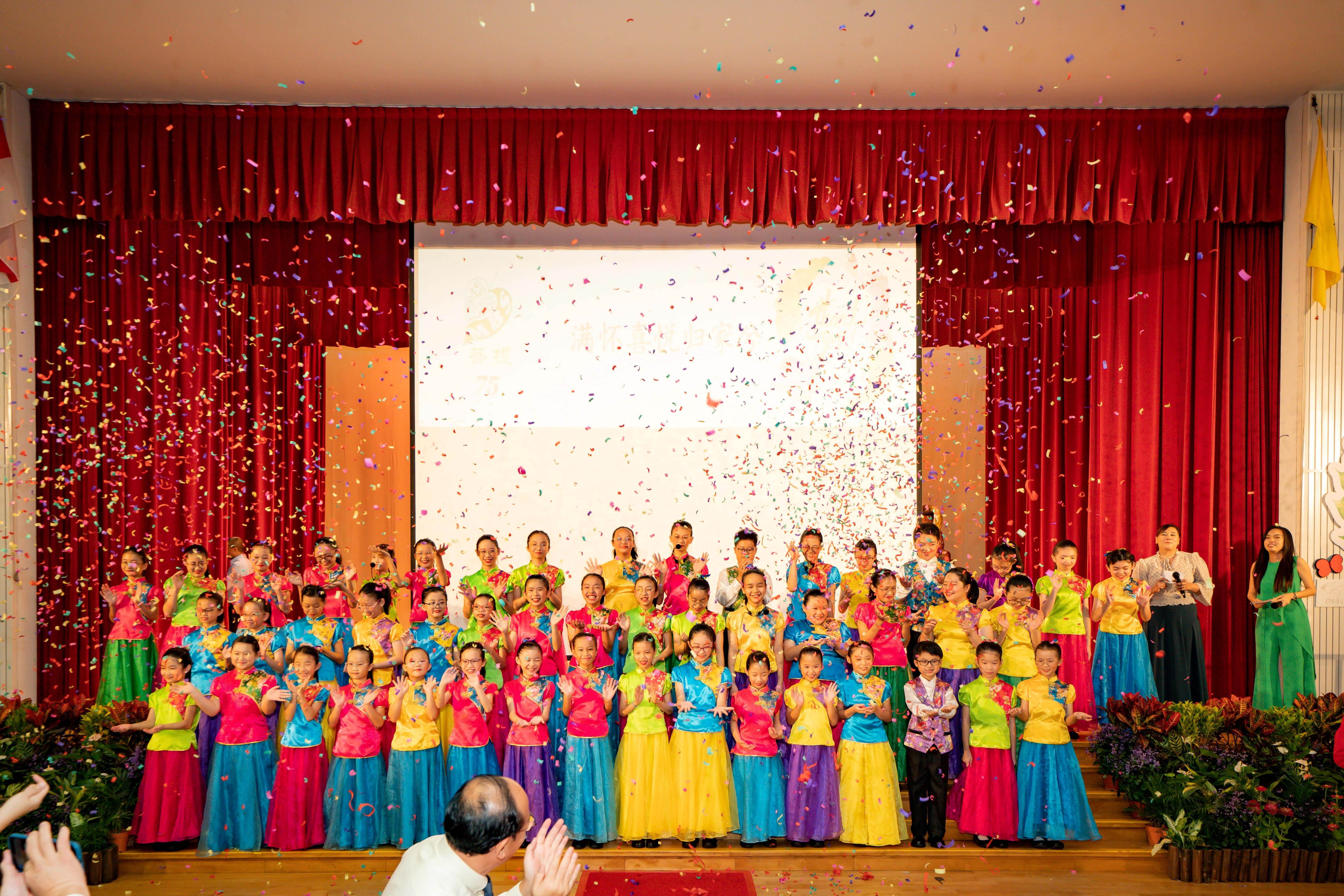 Children in colorful traditional costumes perform on a stage with red curtains as confetti falls around them.
